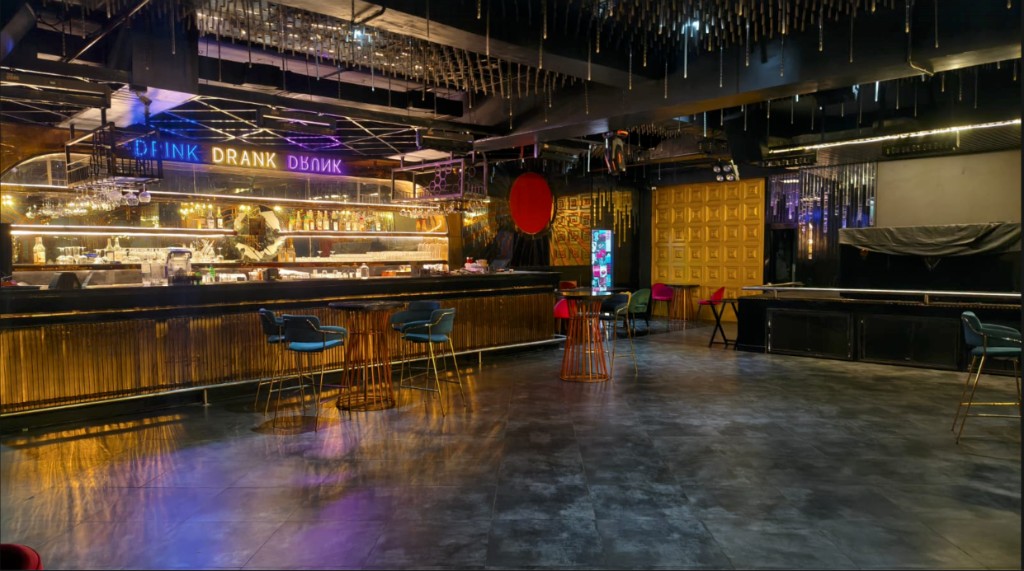 Main bar with a neon Drink Drank Drunk sign, gold ribbed front, and teal bar stools